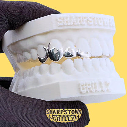 High-polish silver flame drip open frame teeth grillz displayed on dental mold with yellow backdrop.
