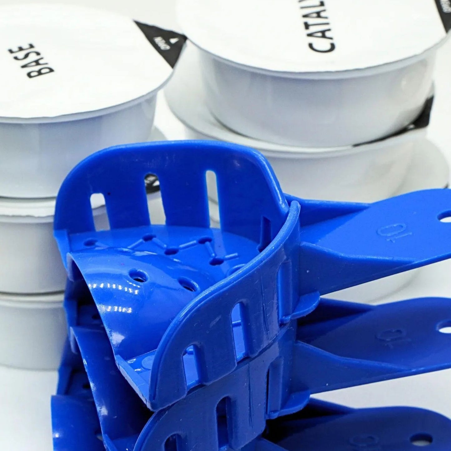 A close-up of several bright blue plastic measuring cups or scoop-like utensils stacked together, with white round containers in the background. The background containers have black text including the word 'BASE' and partial 'CATAL'.